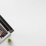 A person typing on a laptop with a notepad and a plant beside it