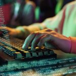 Close-up of hands typing on an illuminated RGB keyboard with code on screen