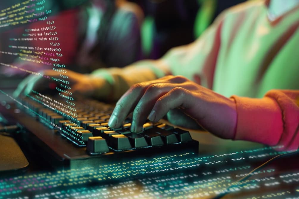 Close-up of hands typing on an illuminated RGB keyboard with code on screen